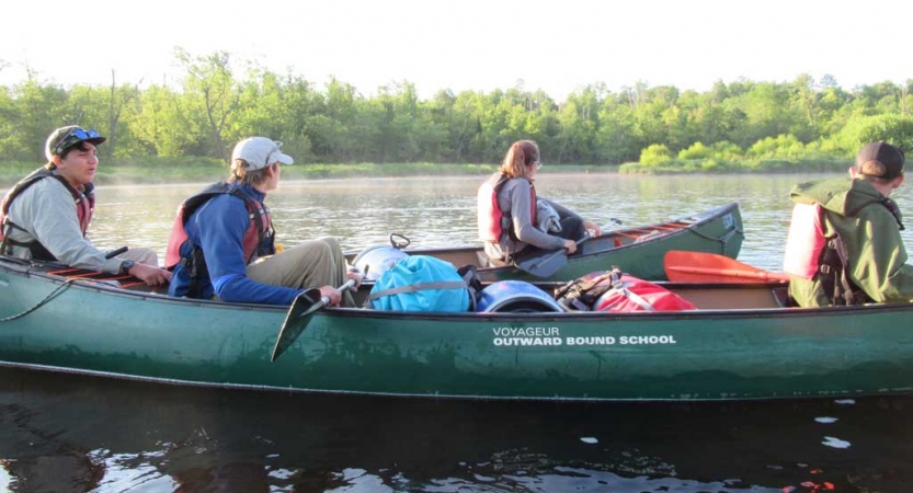 People wearing life jackets take a break from paddling as they sit in canoes on calm water. There is a tree-lined shore in the background. 